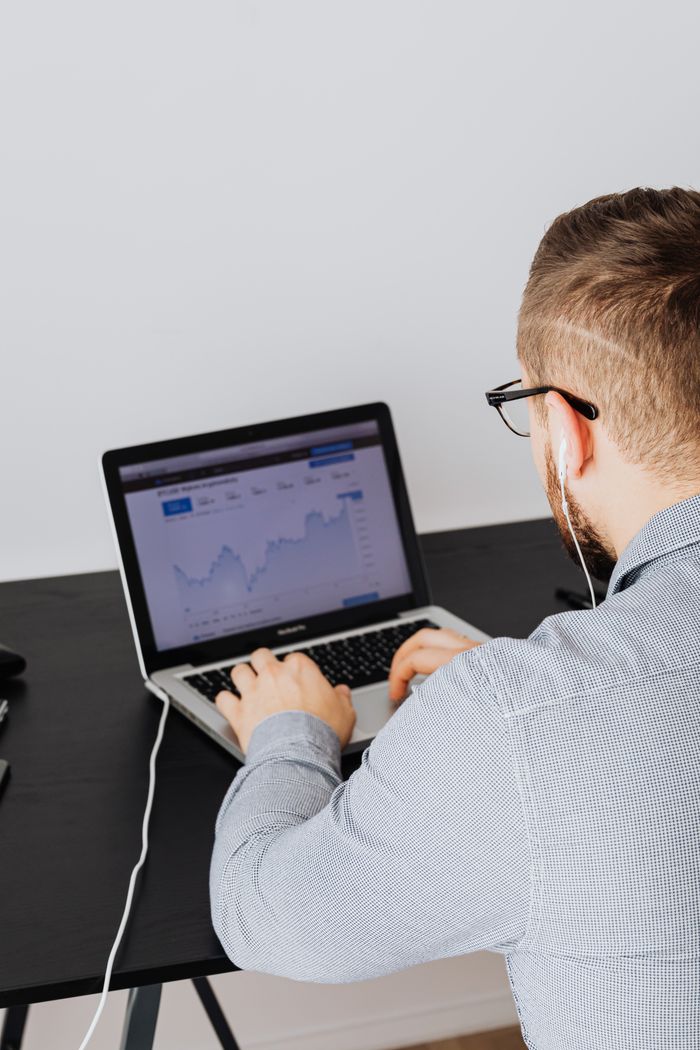 A man is using a laptop indoors, reviewing a stock market trend graph displayed on the screen.